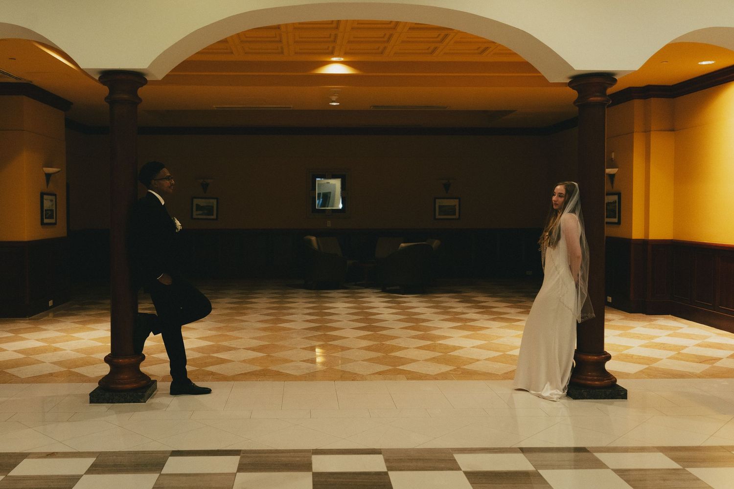 Bride and groom stand apart on checkered floor in elegant arched hall with warm lighting.