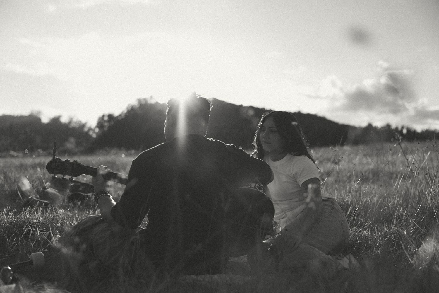 Black and white photo of a couple sitting in a sunlit field, sharing a romantic moment at sunset.