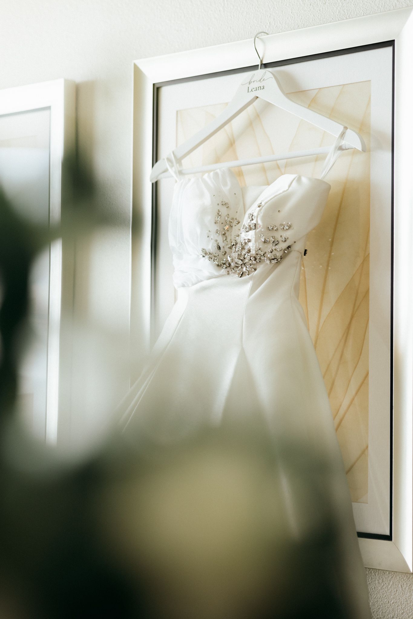 White strapless wedding dress with floral beading hanging on a decorative hanger against a framed wall.