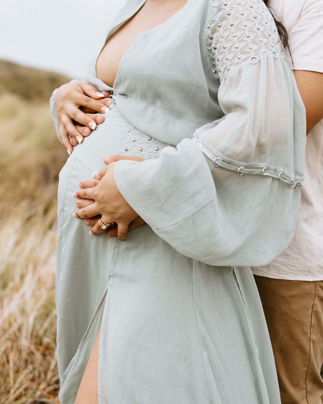 Pregnant woman in sage green dress cradling baby bump outdoors, partner embracing her from behind in a tender maternity photo.