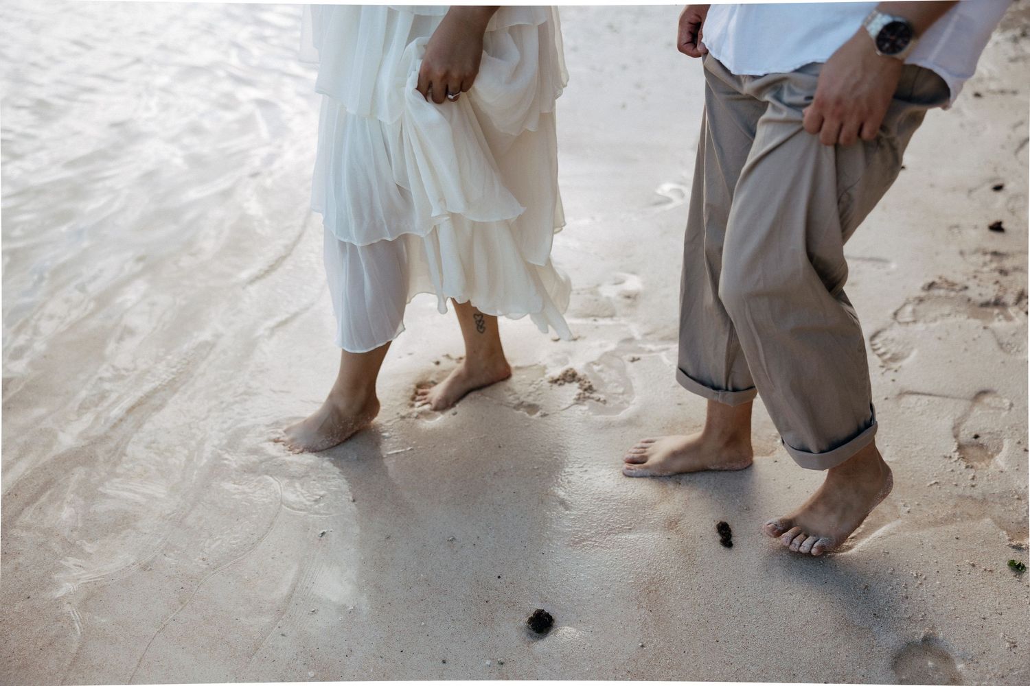 Two barefoot people walking on a sandy beach, one wearing a white dress and the other gray pants.