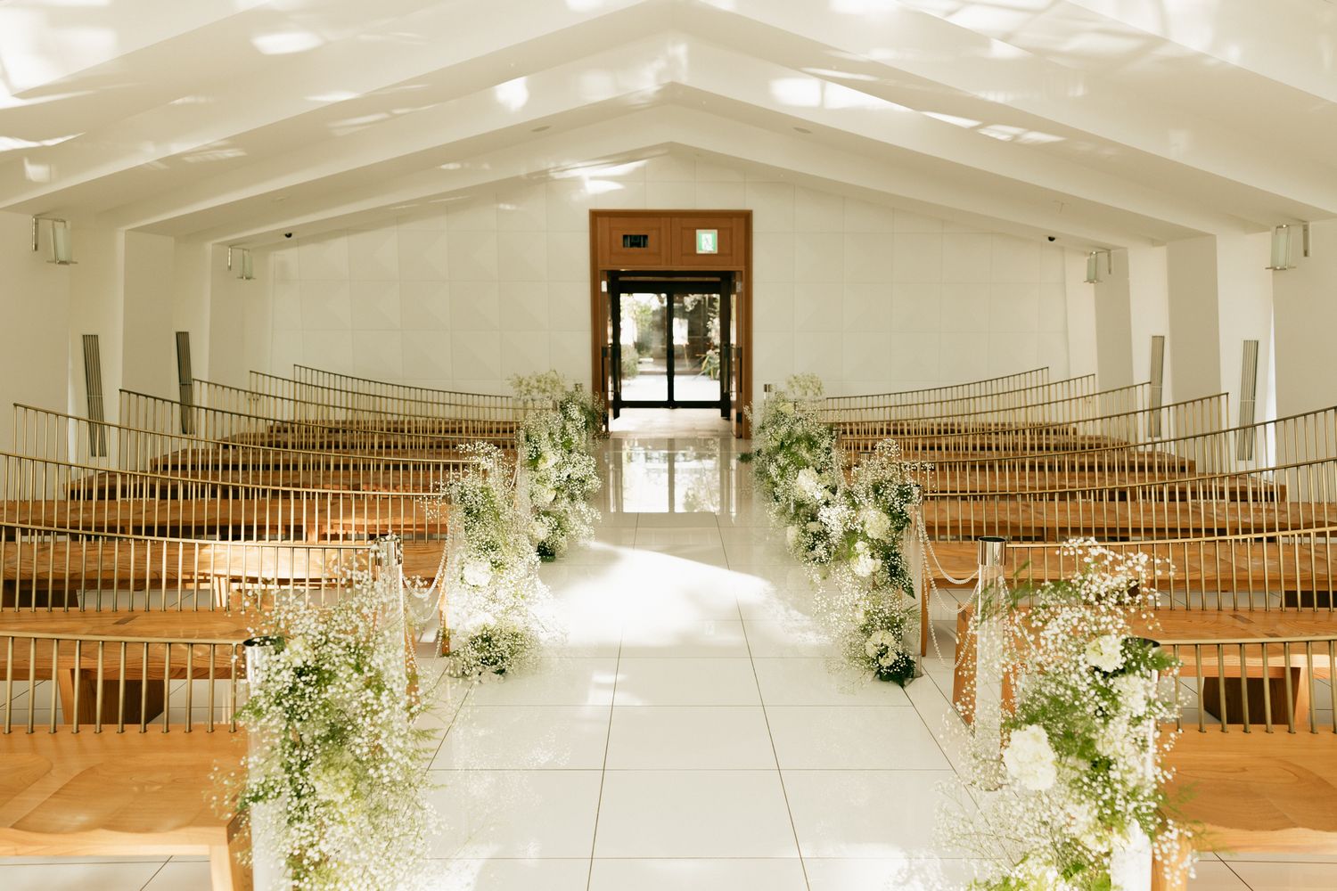 Elegant wedding ceremony aisle with white floral arrangements on wooden pews in a bright, modern chapel.