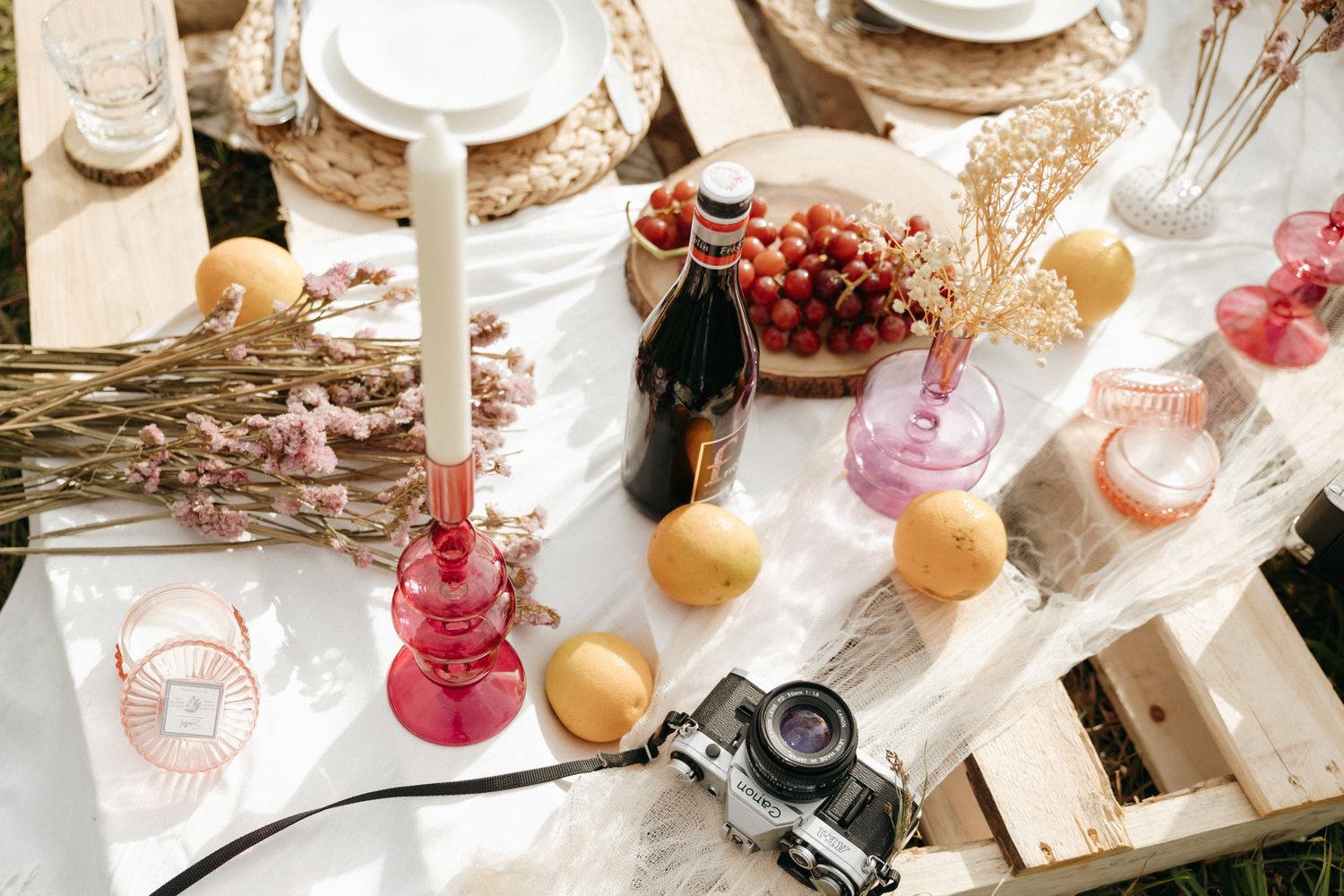 Rustic outdoor picnic table with grapes, peaches, wine bottle, pink glassware, dried flowers, and a vintage camera.