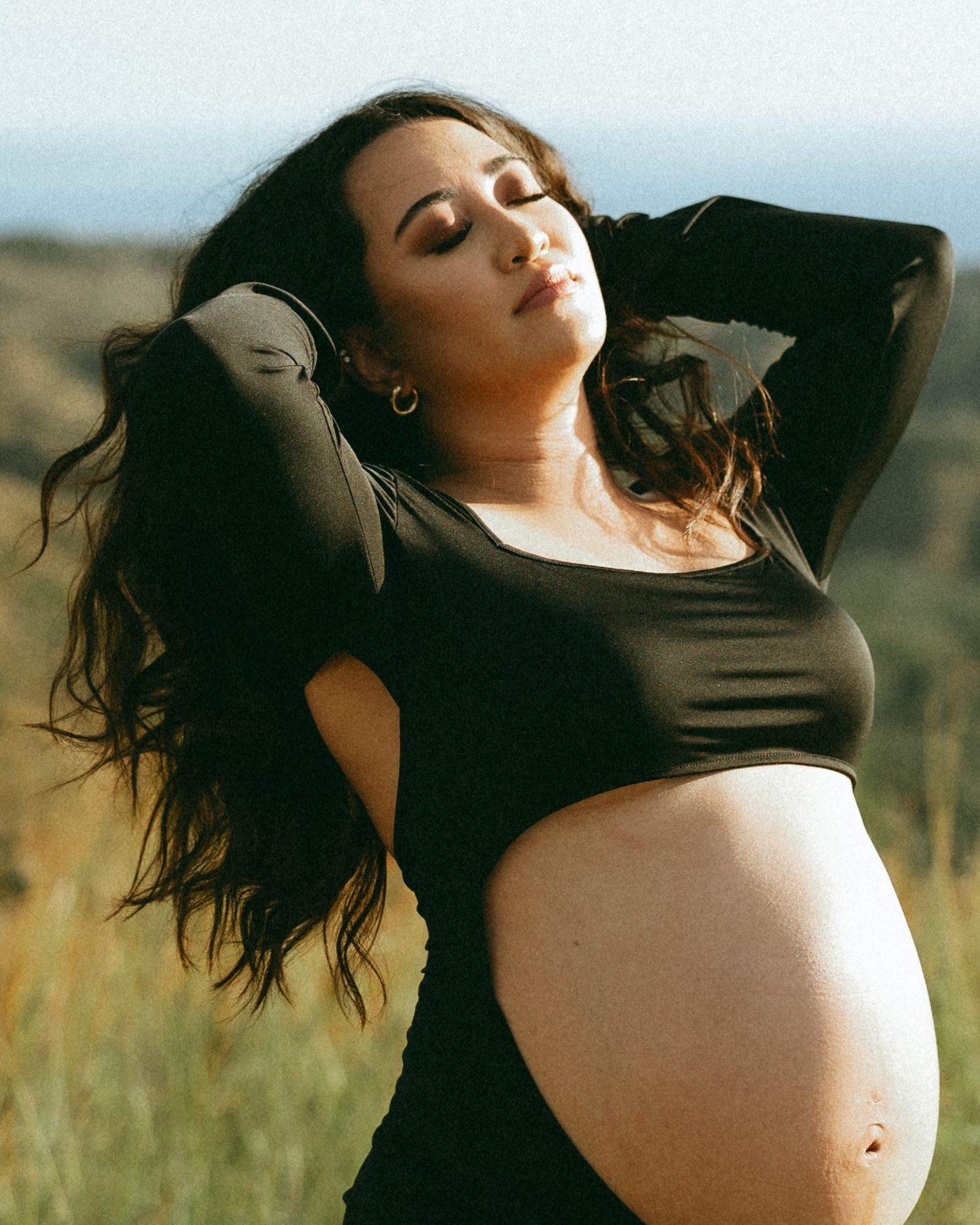 Pregnant woman in black crop top poses outdoors in golden field, hands behind head, eyes closed, with flowing dark hair.