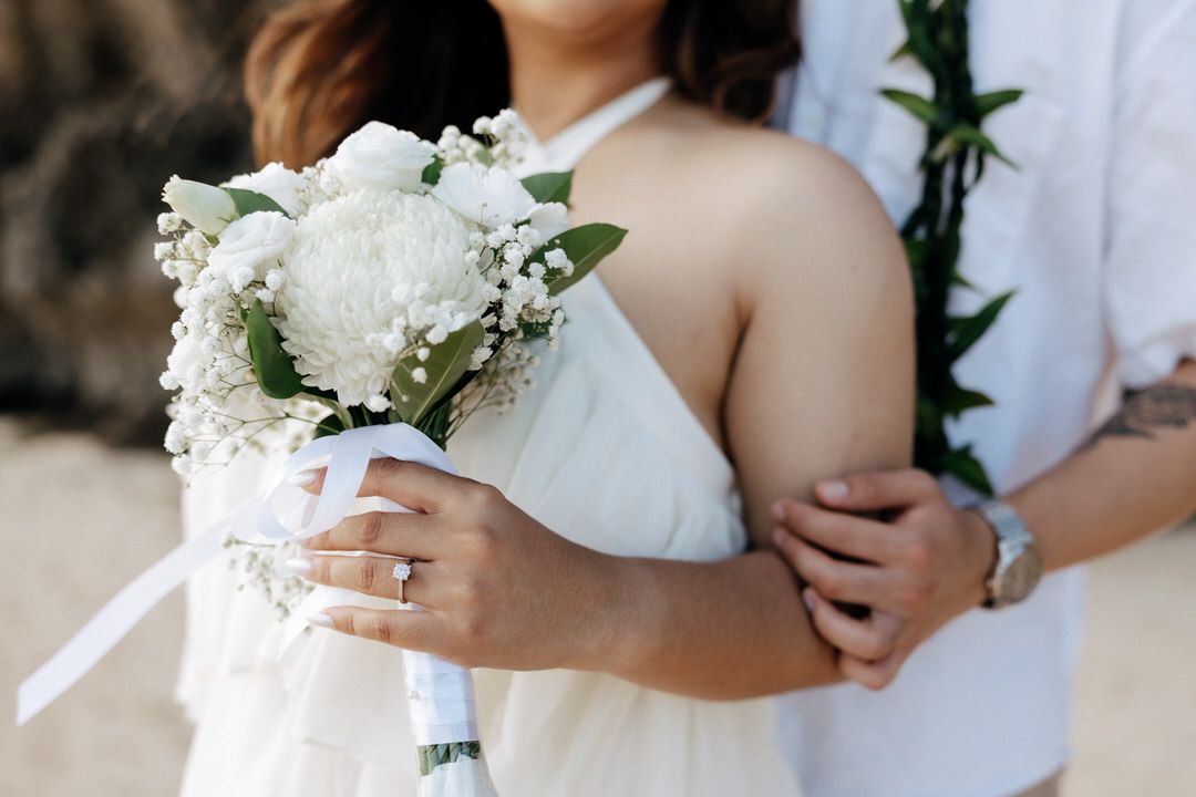 Bride holding white floral bouquet with roses and baby's breath while groom embraces her at beach wedding.
