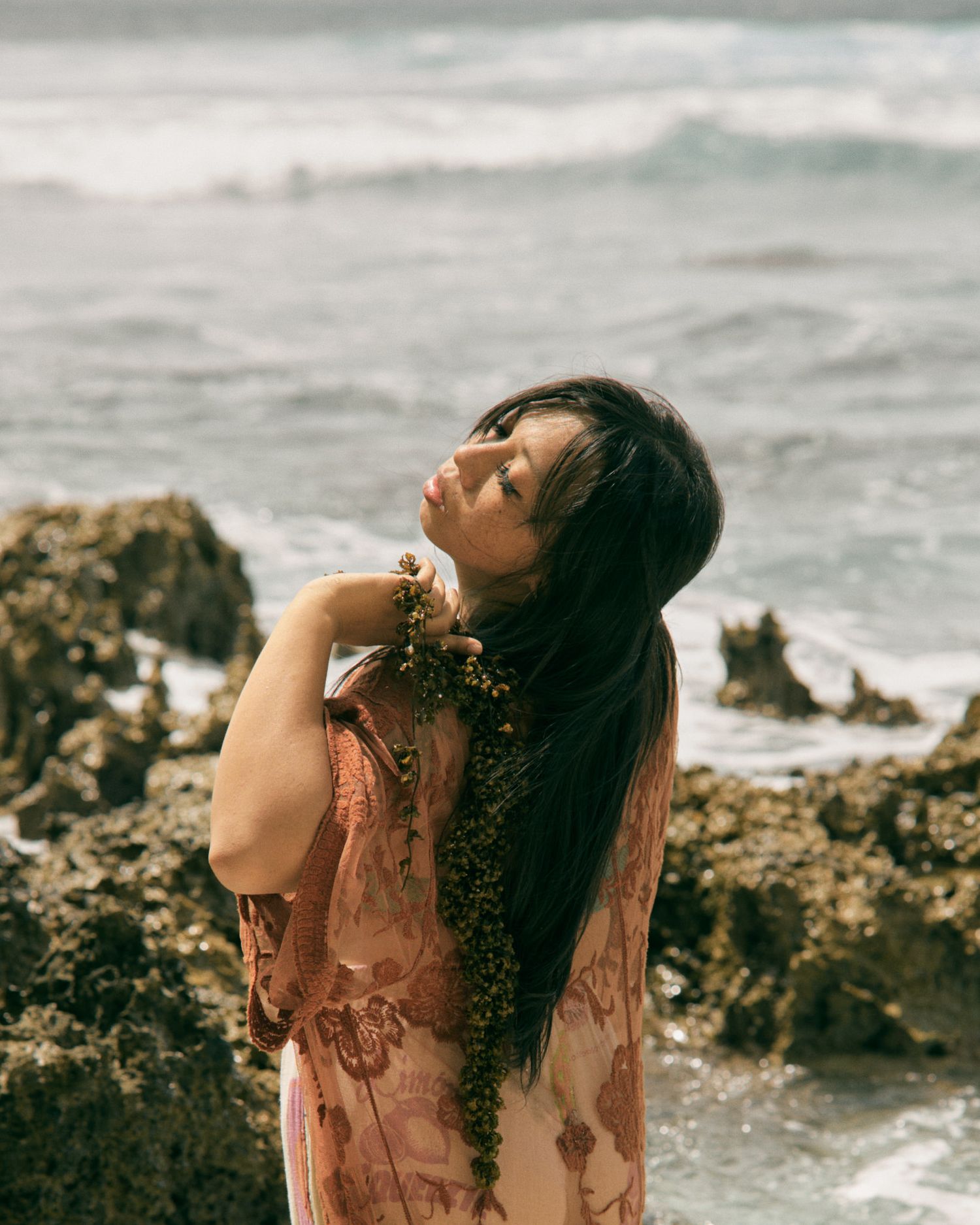 A young woman in a floral dress holds seaweed while posing on rocky coastal cliffs with ocean waves behind her.