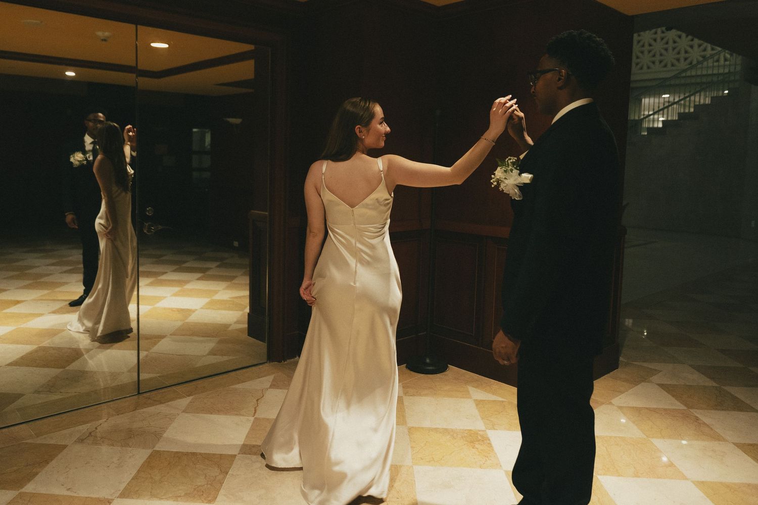Bride in a white satin gown and groom in black suit dancing near a mirror in a dimly lit venue.
