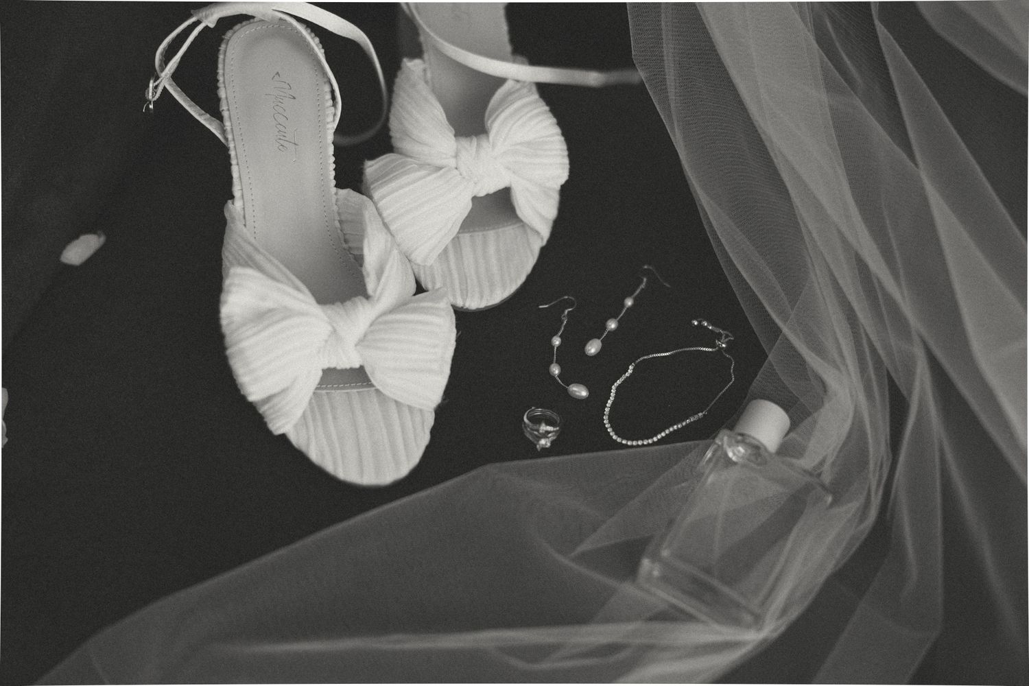 Black and white flat lay of white bow-detail bridal heels, jewelry, and tulle wedding veil.