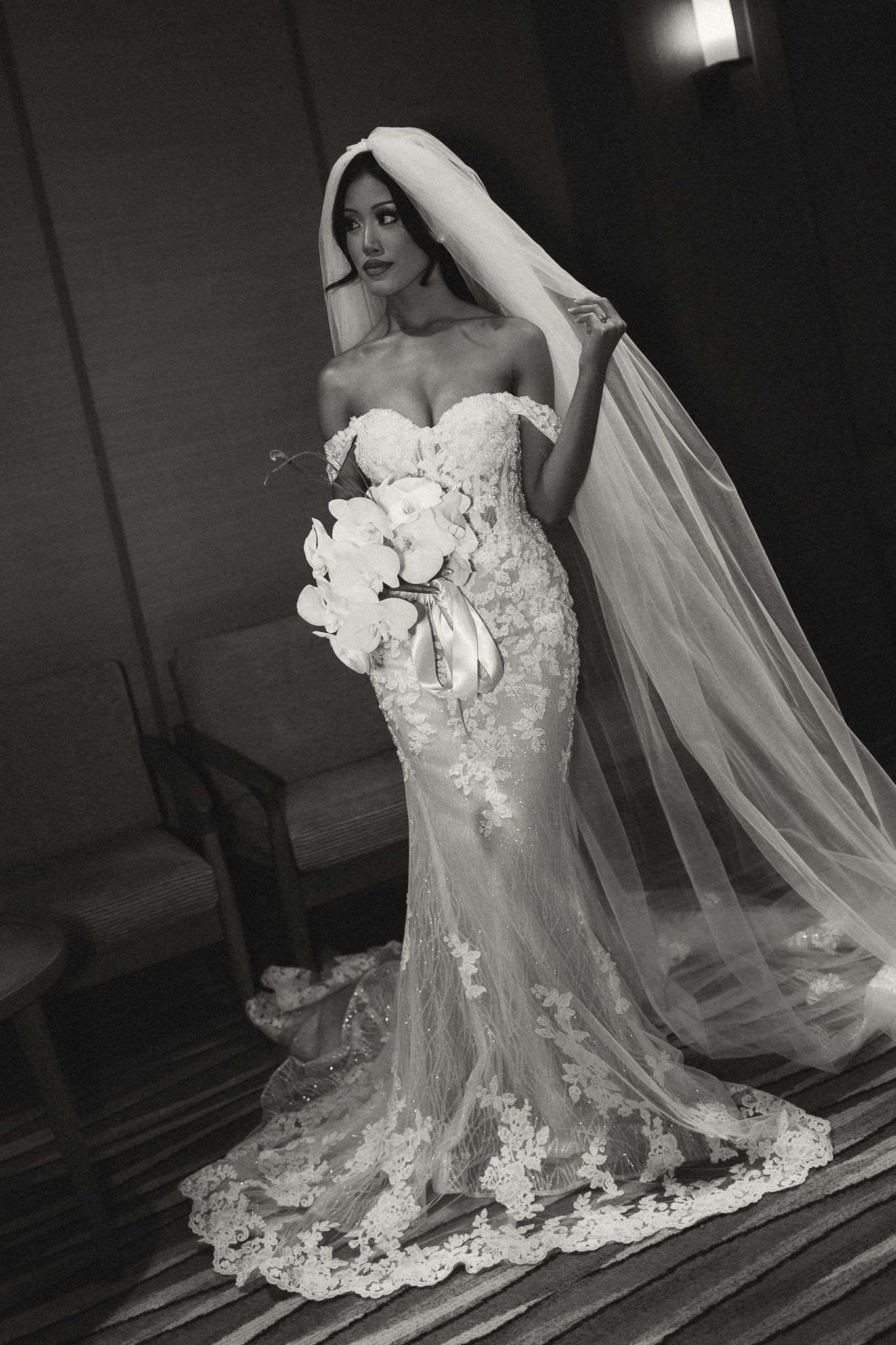 Bride in lace mermaid wedding gown with off-shoulder neckline, holding white bouquet, wearing long veil, black and white photo.