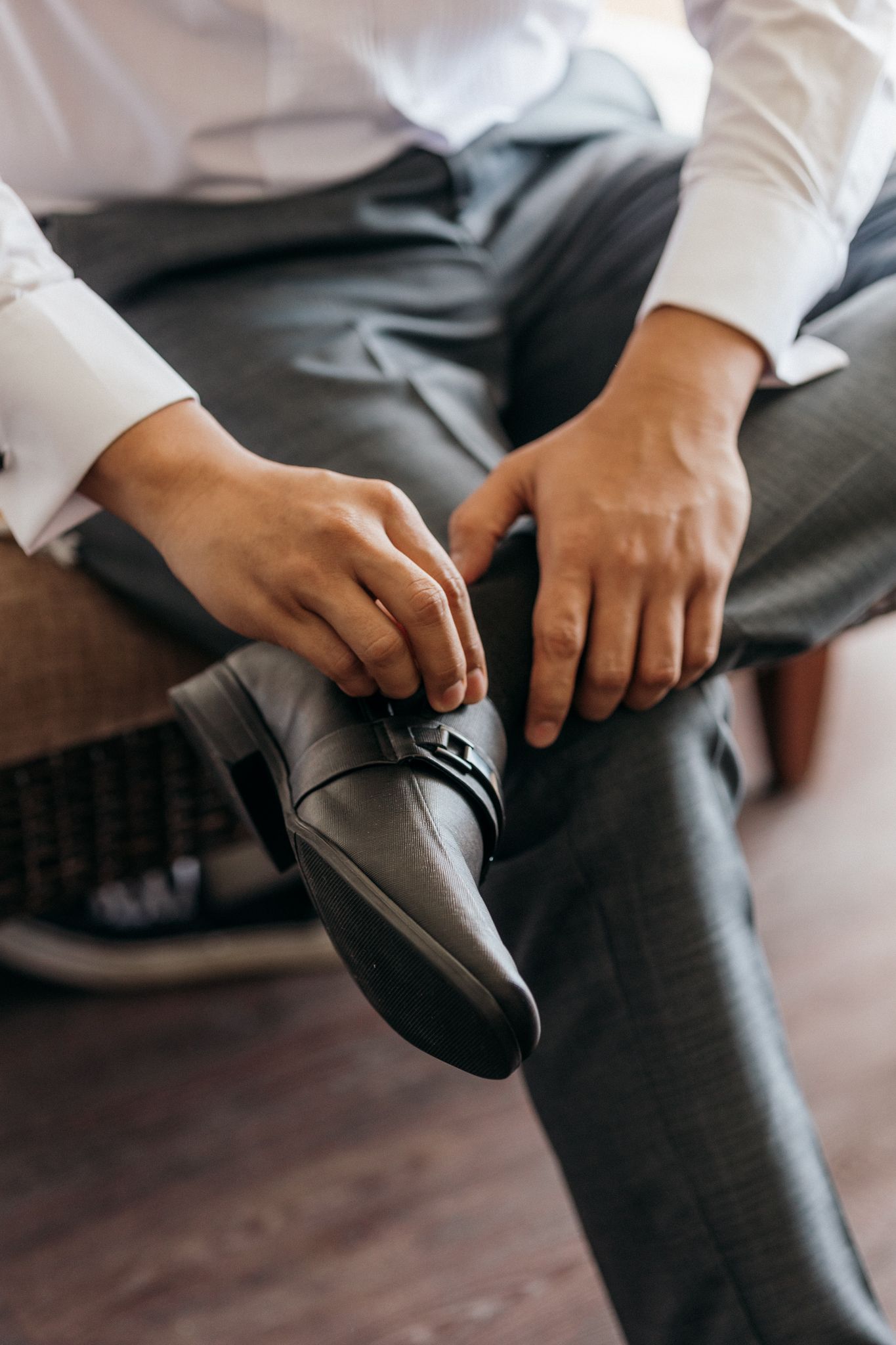 Man in white dress shirt and gray trousers adjusting black leather dress shoes while seated.