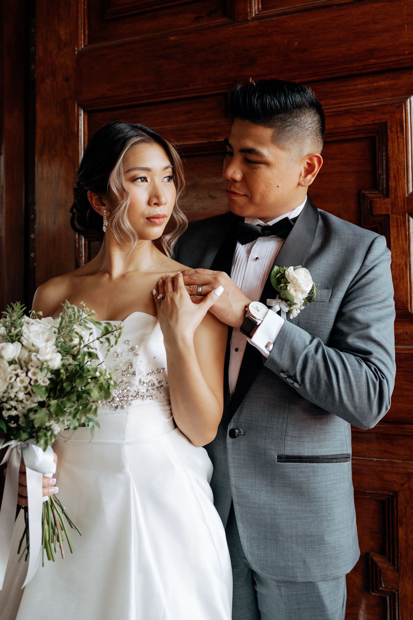 Groom in gray suit placing ring on bride in white gown holding bouquet, posing before wooden doors.