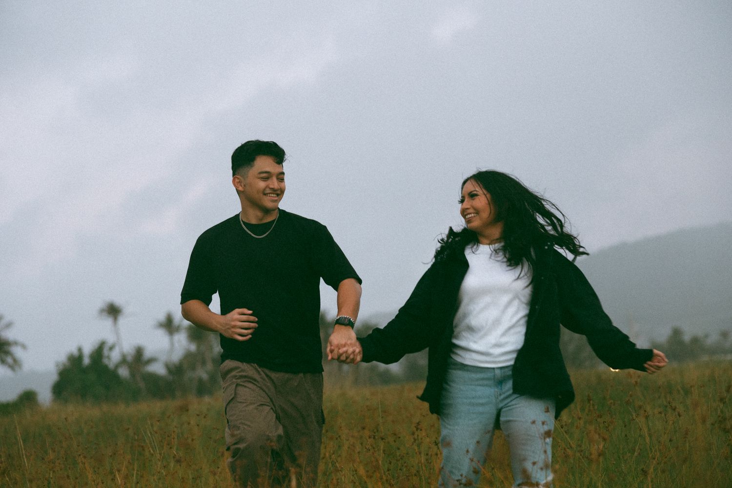 A happy Asian couple holds hands and runs through a grassy field under a cloudy sky.
