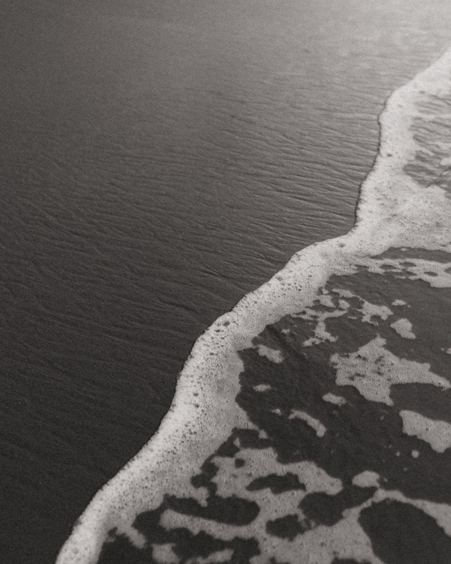 Black and white aerial view of ocean waves washing onto a sandy beach with foamy surf.