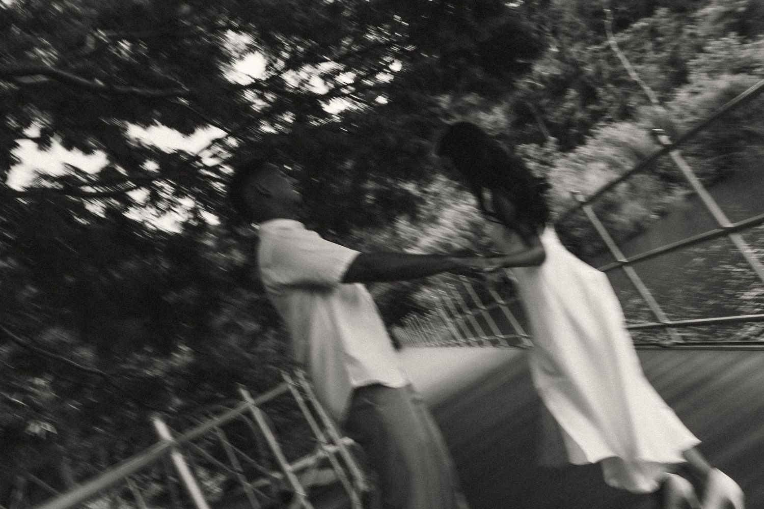 Black and white photo of two people dancing outdoors near trees and a metal railing.