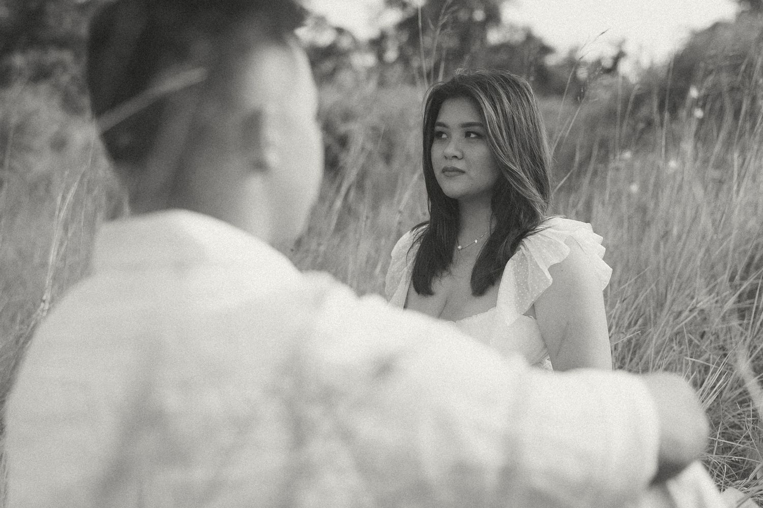 Black and white photo of a young woman in a white dress sitting in tall grass, gazing at someone in the foreground.
