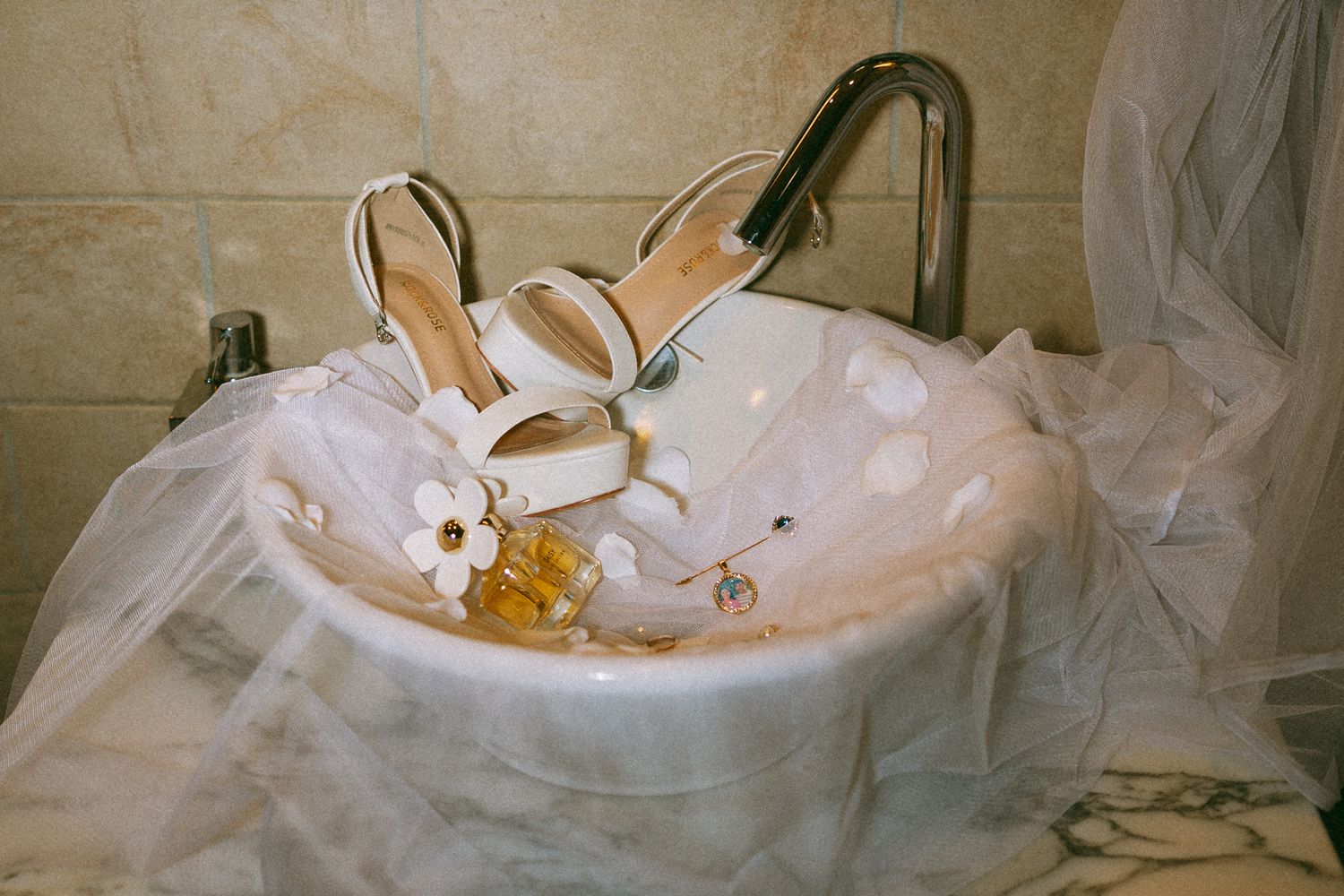 White strappy bridal heels with floral detail resting in a marble sink with tulle veil and perfume.