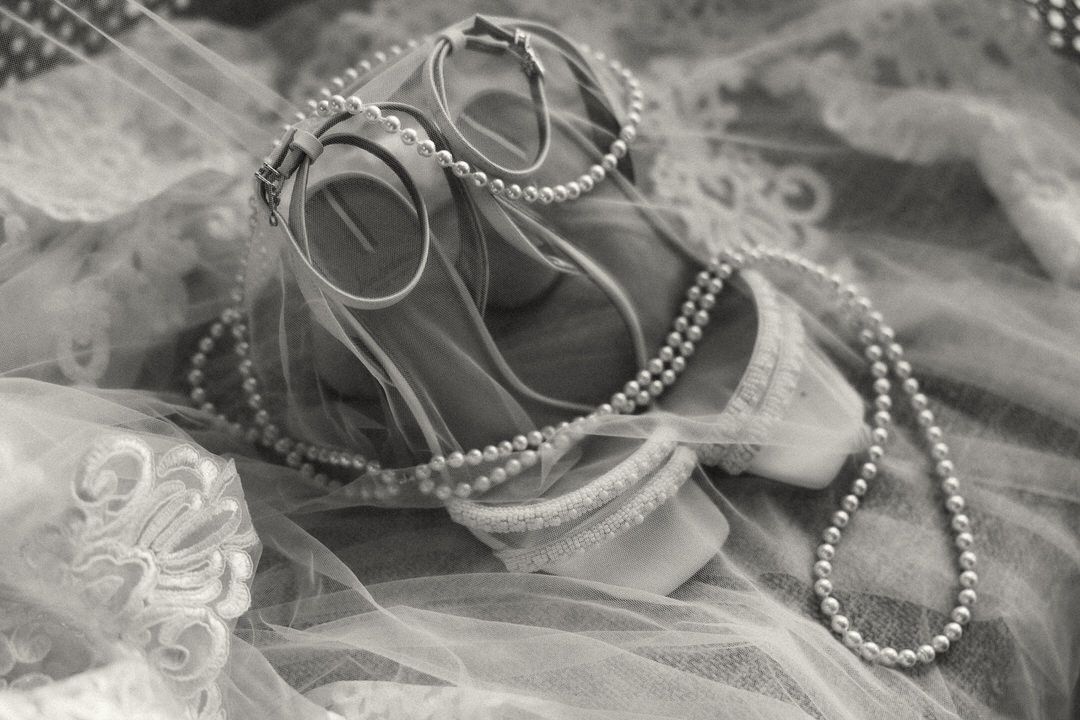 Black and white photo of elegant bridal heels with pearl necklace on lace veil fabric.