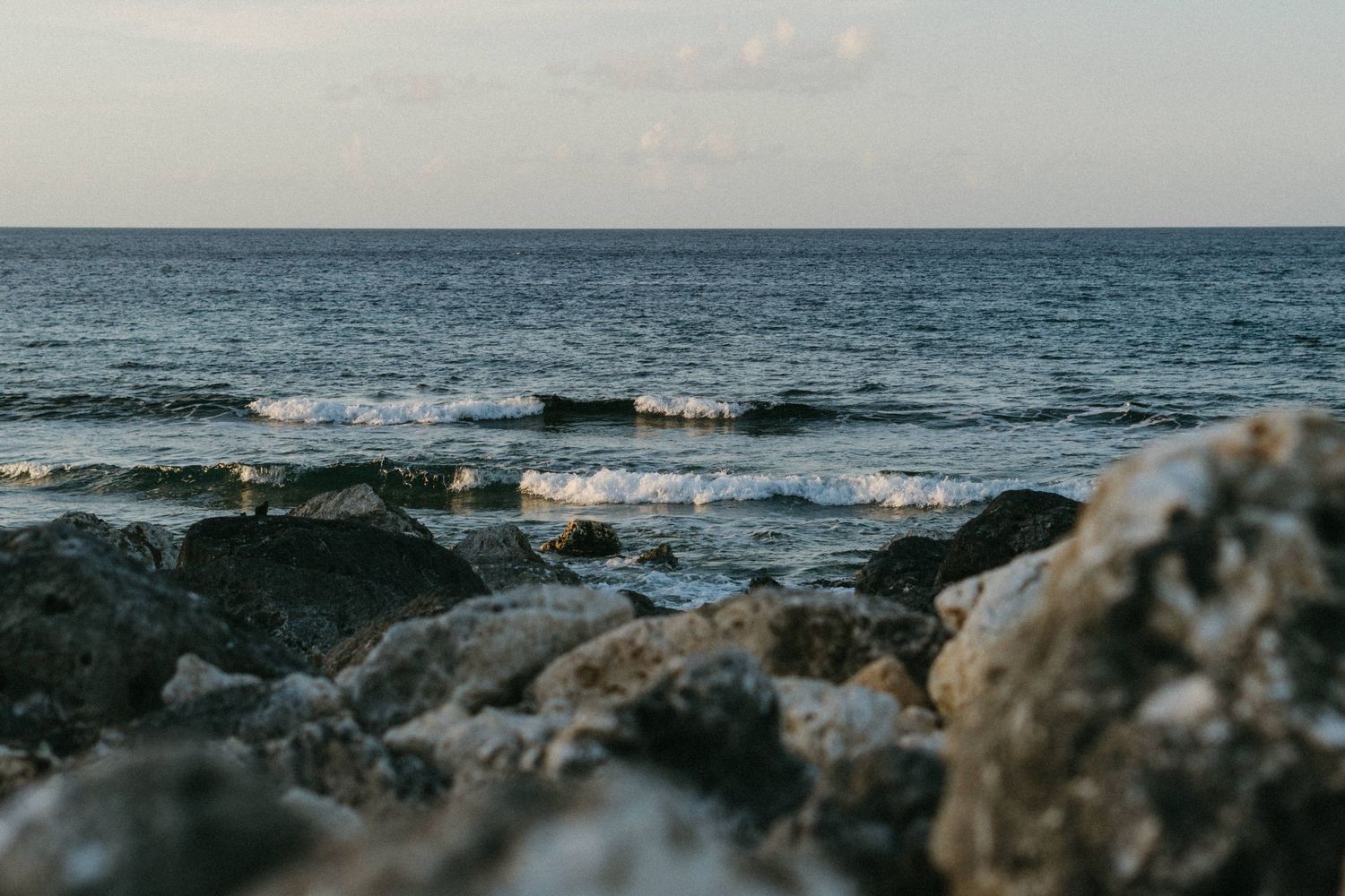 Rocky shoreline with gentle ocean waves and a calm horizon under a cloudy sky.