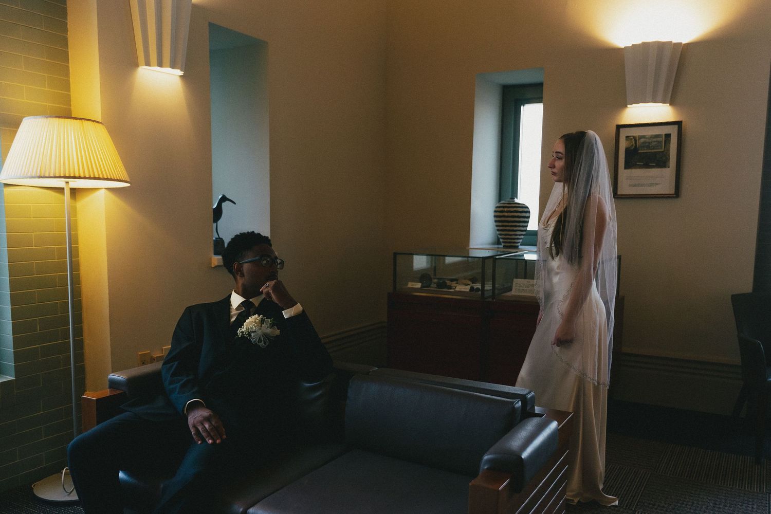 Bride in white gown and veil stands near groom in dark suit seated on leather sofa in warmly lit elegant room.