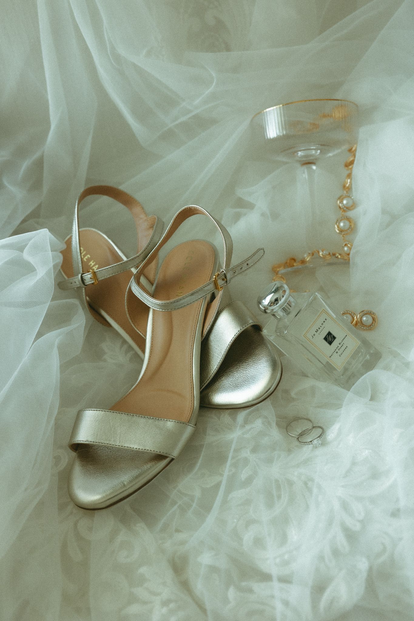 Gold strappy heeled sandals with perfume, pearl jewelry, and champagne glass on white bridal tulle fabric.