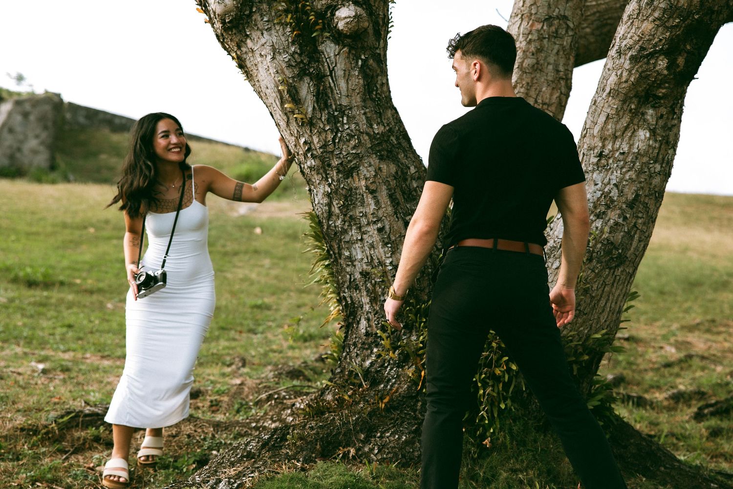 A woman in a white dress holds a camera while smiling at a man near a large tree outdoors.