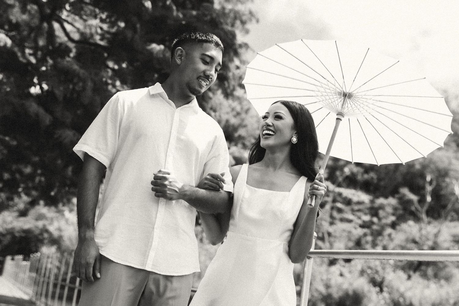 Couple in white attire walking together, woman holding parasol umbrella, smiling outdoors in black and white photo.