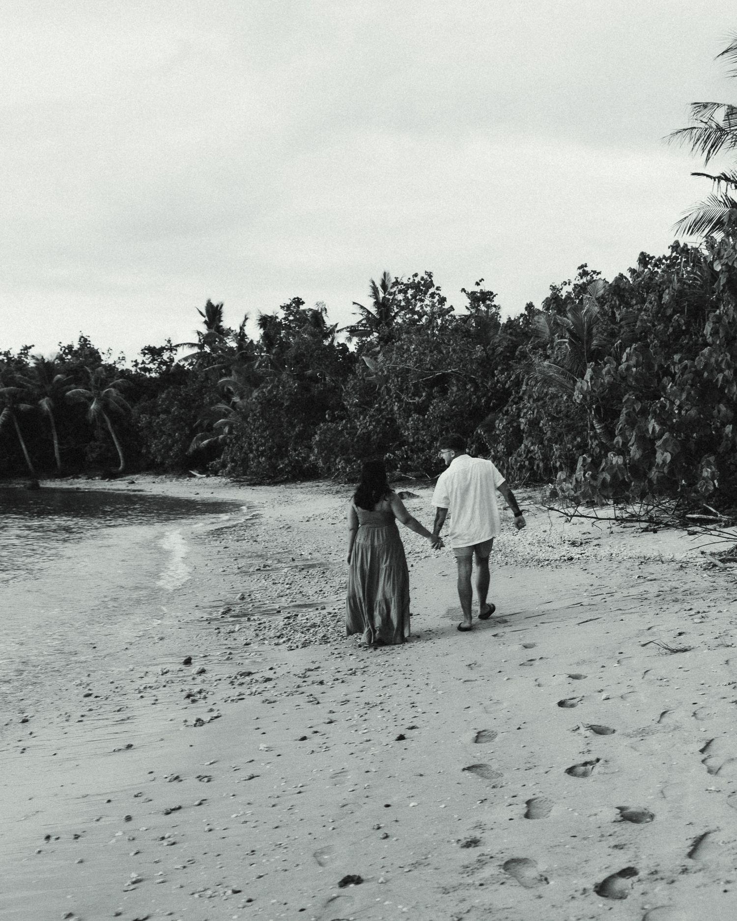Black and white photo of a couple holding hands walking on a tropical beach with palm trees.