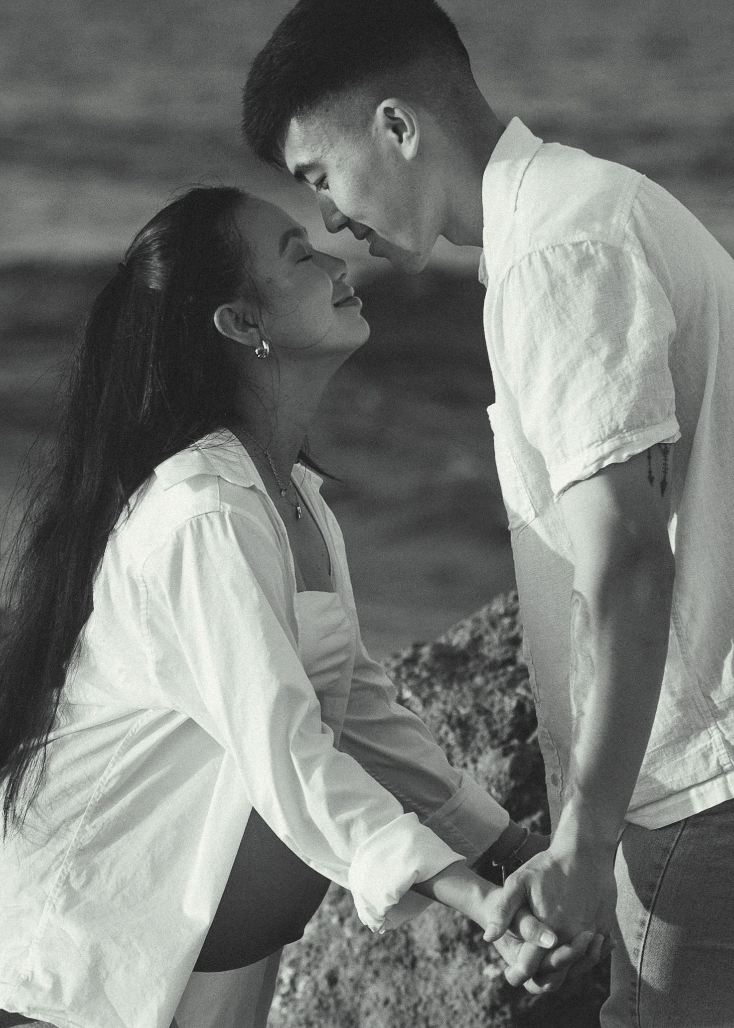Black and white photo of a romantic couple holding hands and leaning in for a kiss near a rocky beach.