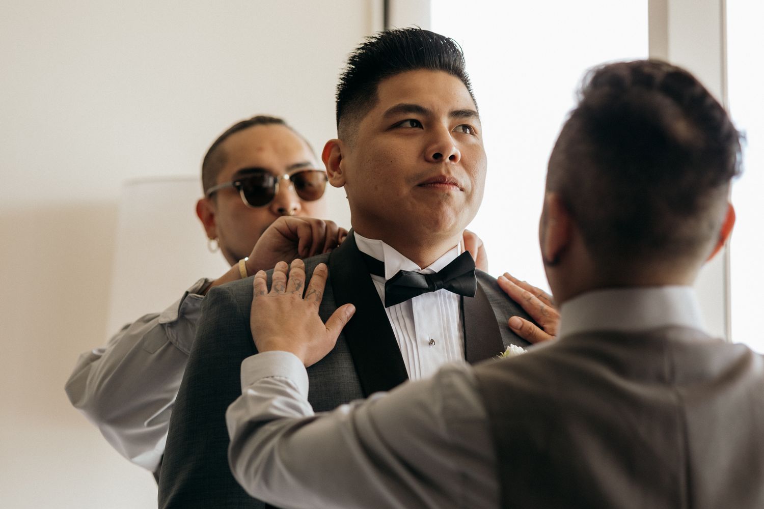 Groomsmen helping groom adjust his tuxedo vest and bow tie while getting ready for wedding ceremony.