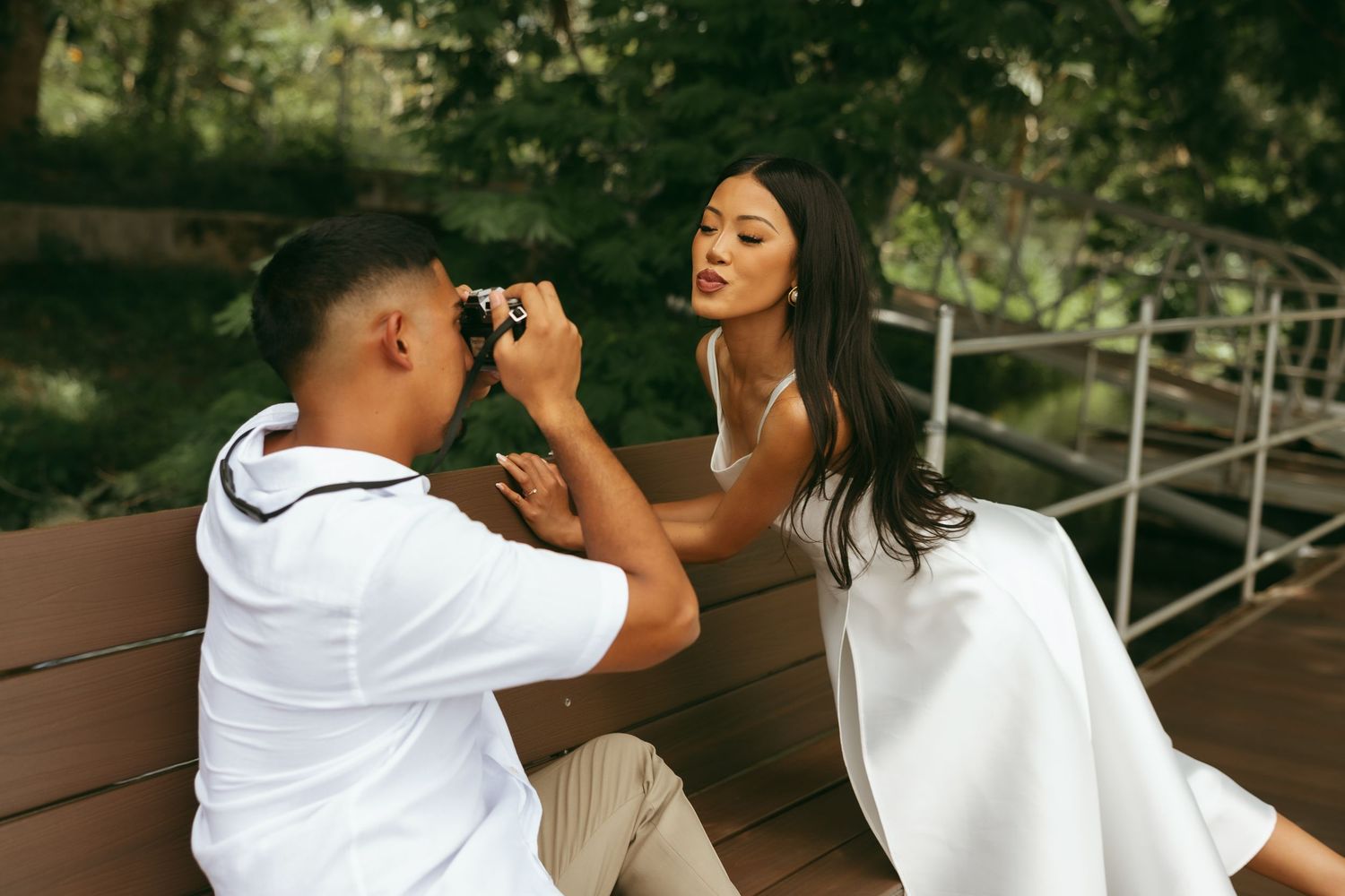 A man in white shirt photographs a woman in white dress sitting on a bench in a lush green park setting.