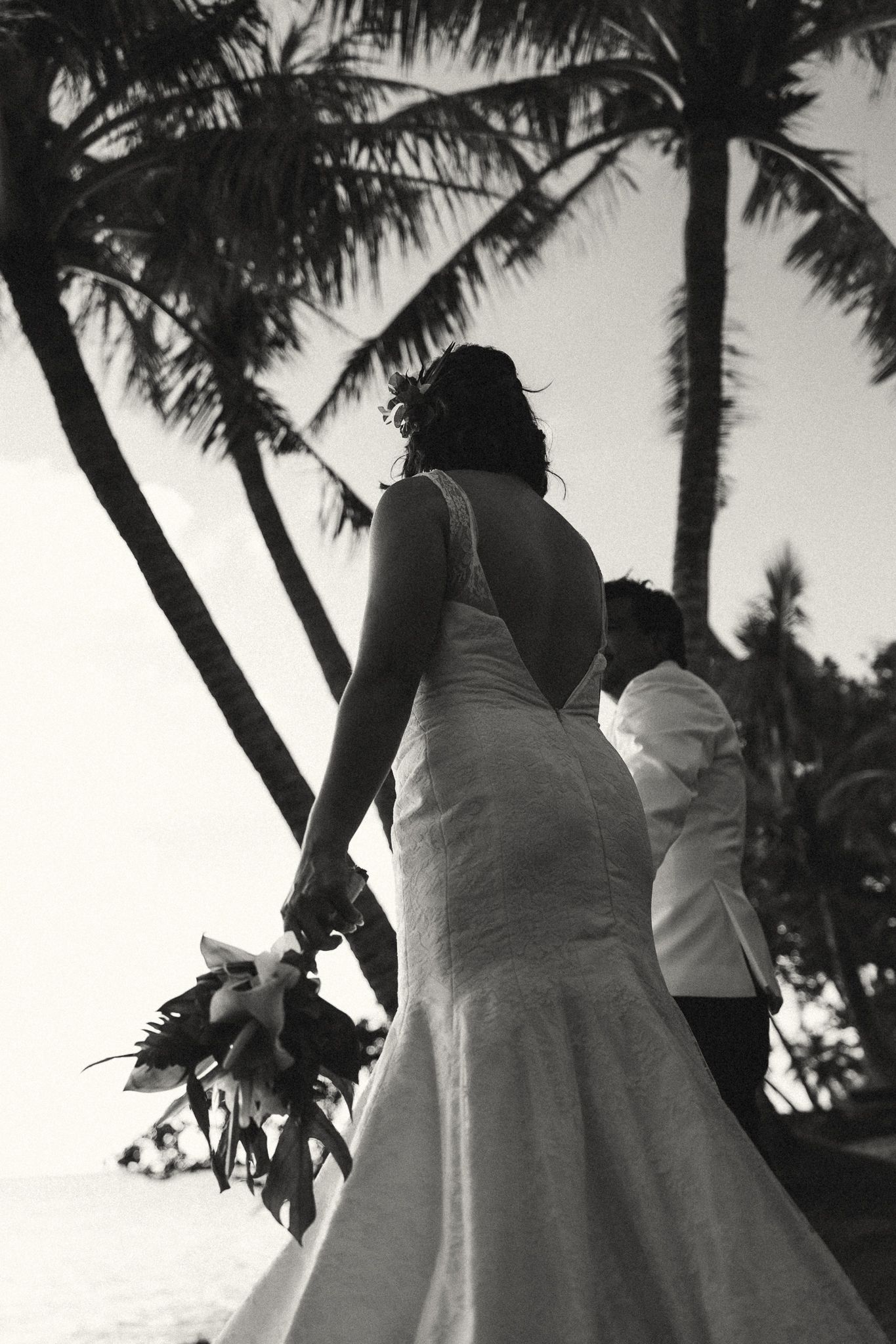 Black and white photo of bride and groom holding hands under palm trees at a tropical beach wedding.
