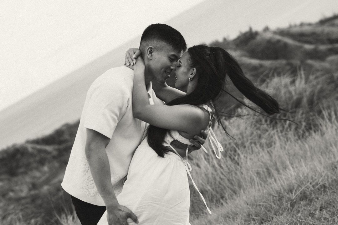 Black and white photo of a couple sharing a romantic kiss outdoors in a grassy coastal landscape.