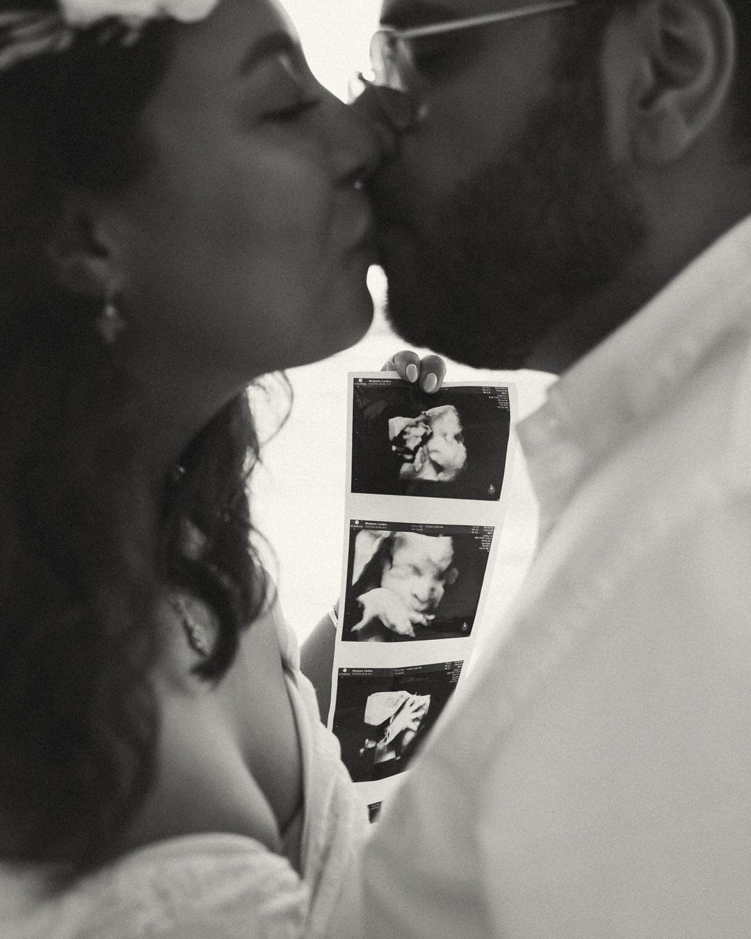 Black and white photo of a couple kissing while holding ultrasound photos, announcing their pregnancy.
