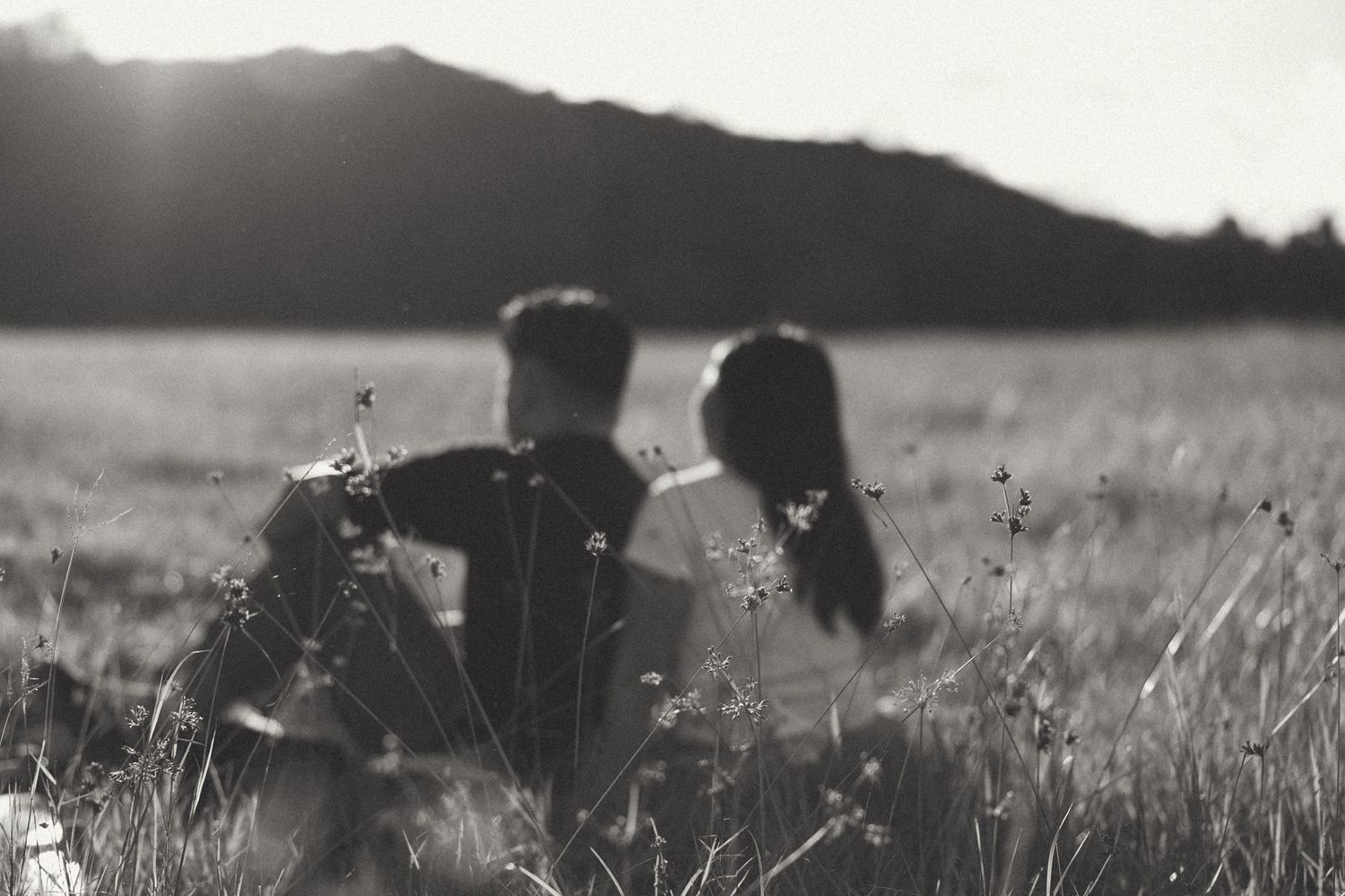 Black and white photo of a couple sitting together in a grassy field with wildflowers and mountains in the background.