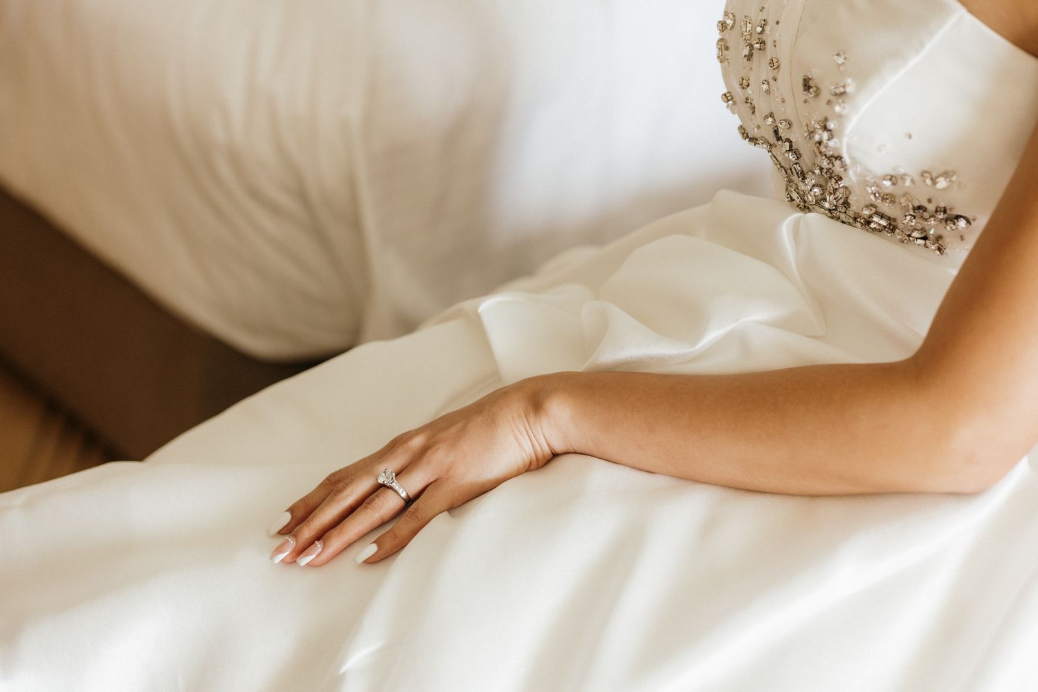Bride in beaded wedding gown resting hand on white fabric, showcasing elegant diamond engagement ring.