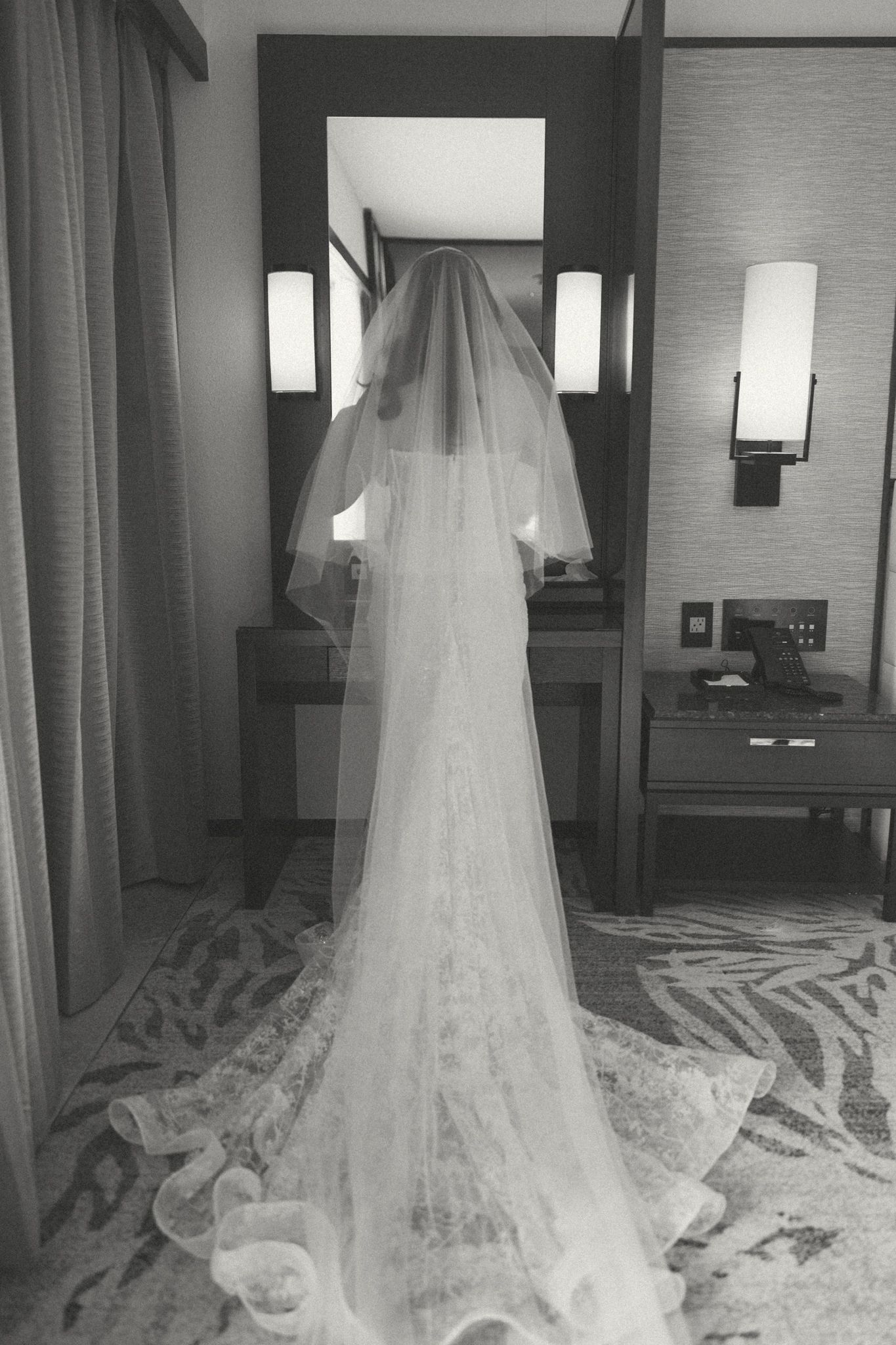 Black and white back view of bride in lace wedding gown with cathedral veil at mirror.