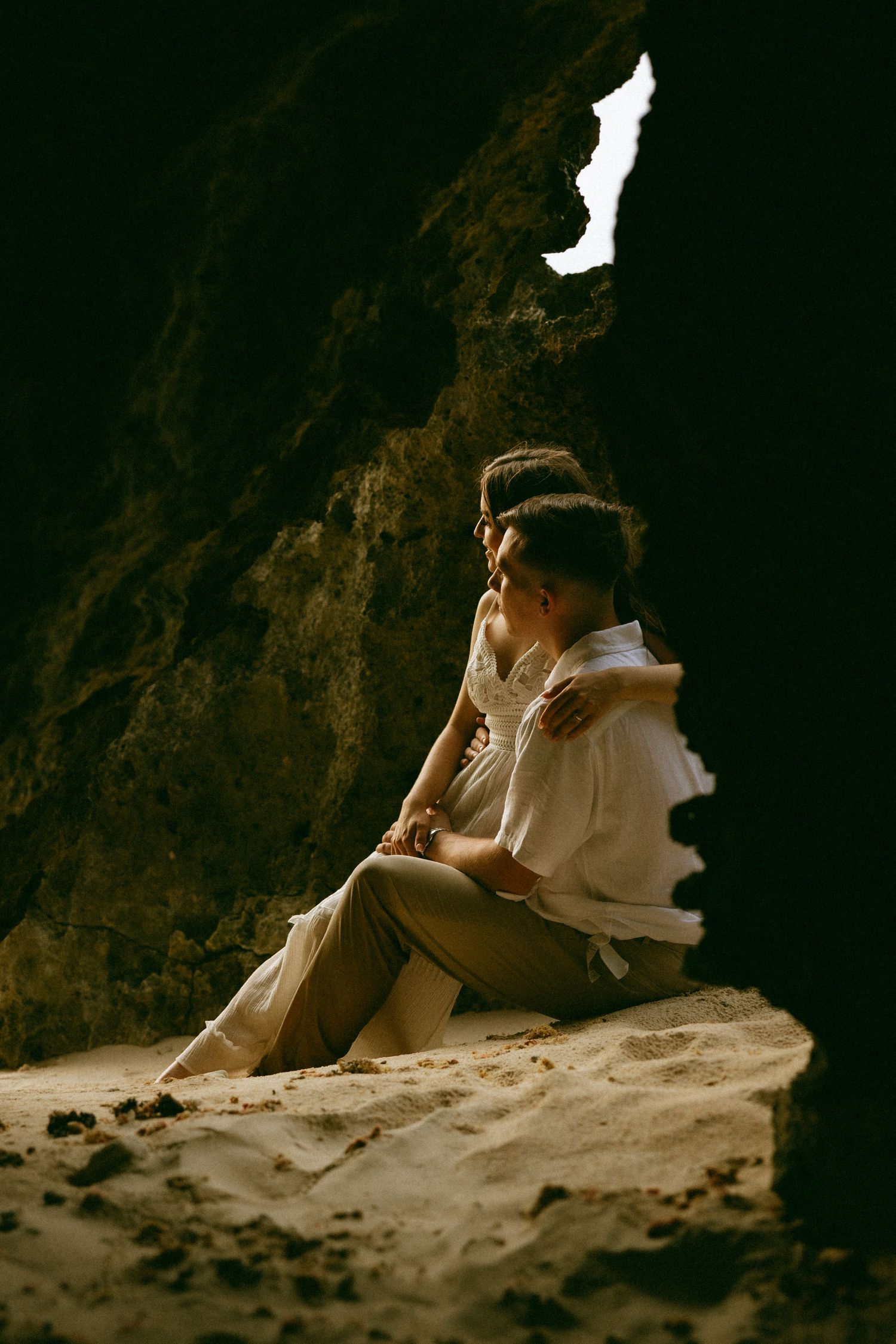 Romantic couple sitting together in a sandy cave alcove, dressed in elegant white and beige attire, sharing an intimate moment.