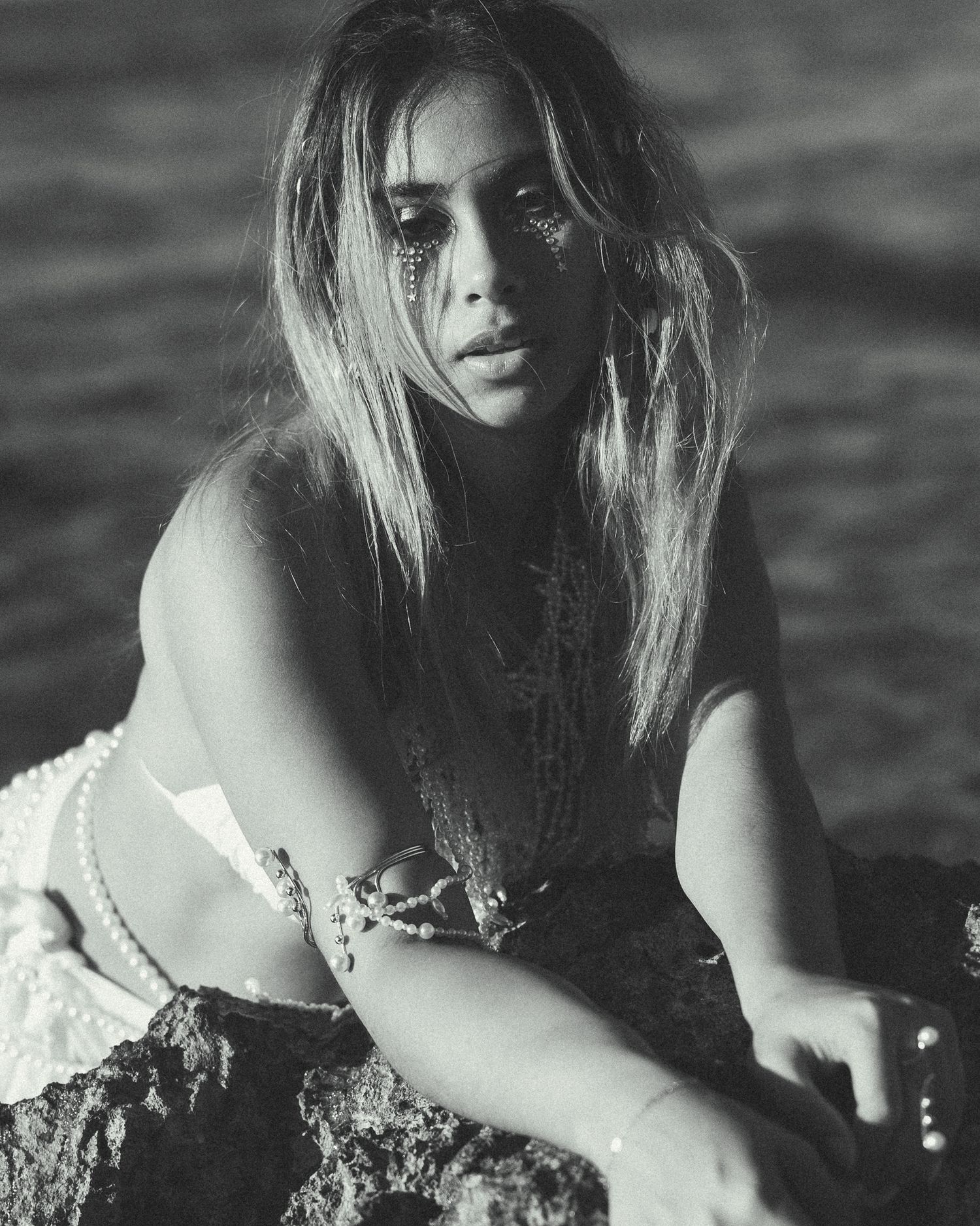 Black and white photo of a young woman with long wavy hair sitting on rocks by the ocean wearing jewelry.