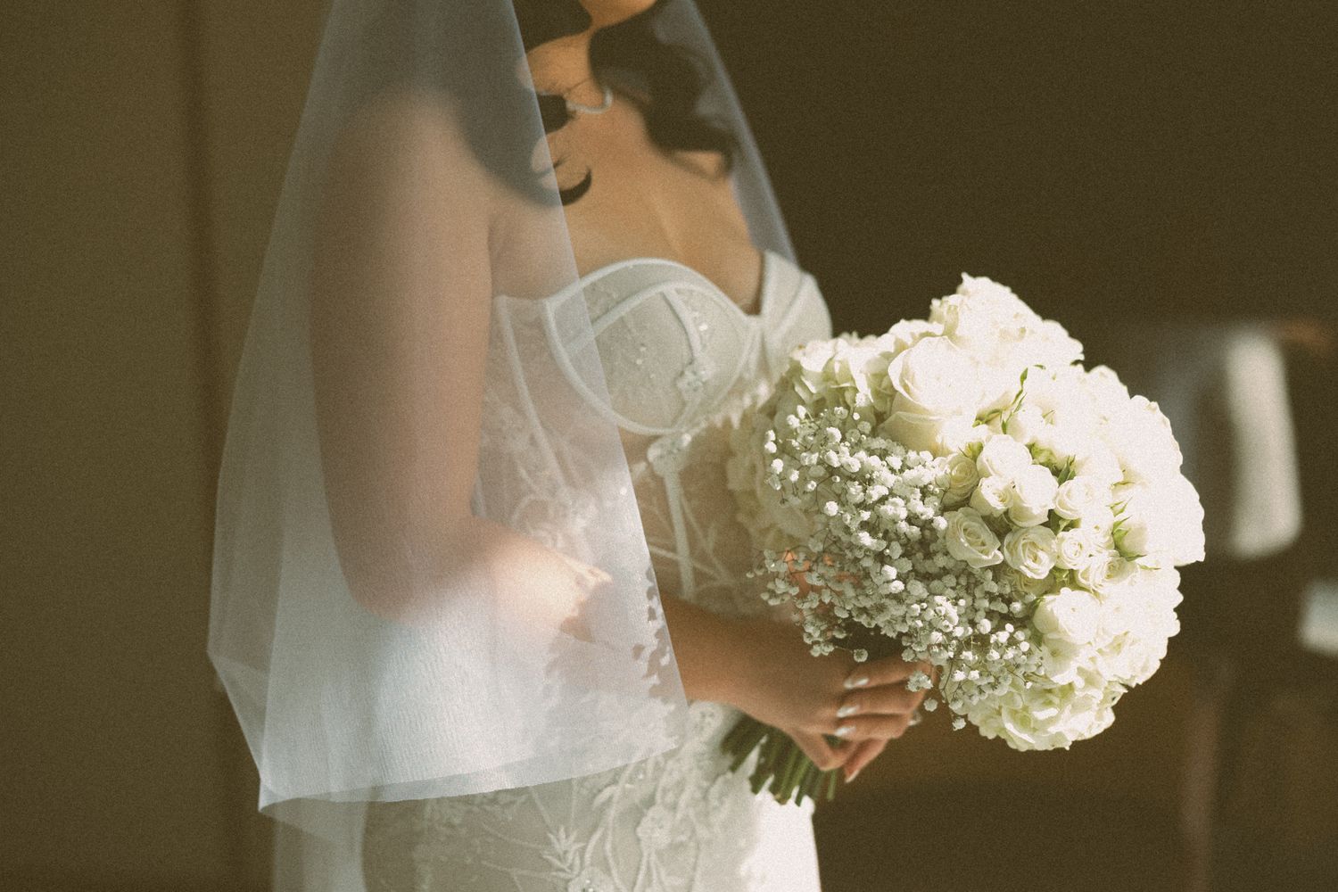 Bride in lace wedding gown and veil holding elegant white rose and baby's breath bouquet on her wedding day.