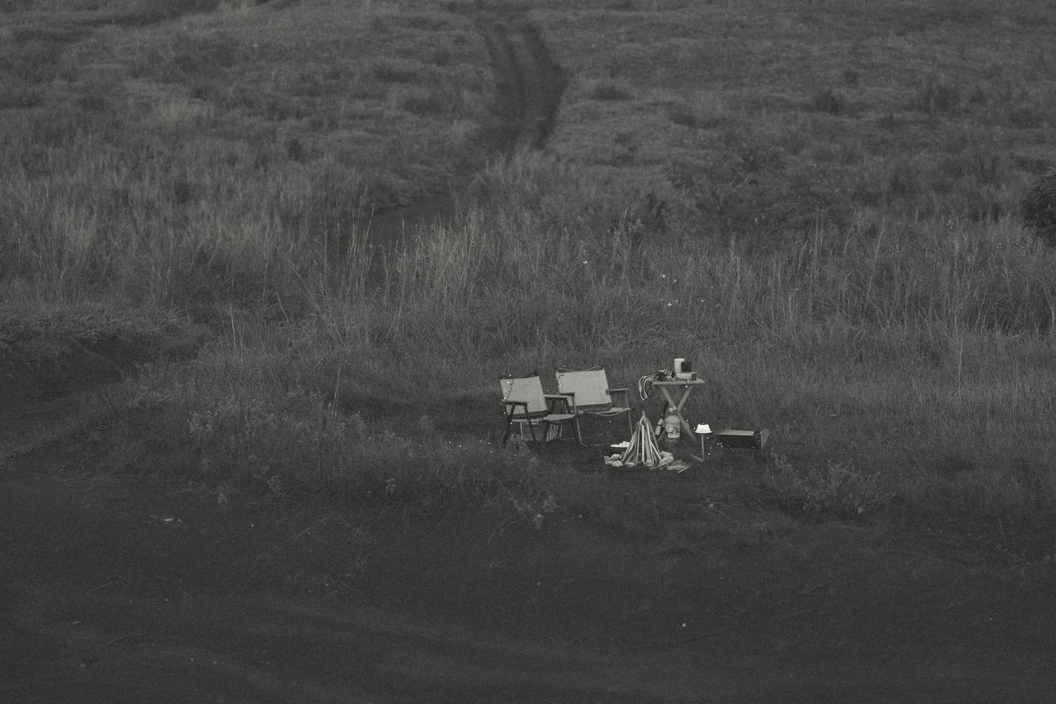Black and white photo of outdoor chairs and equipment in a grassy field with tire tracks visible in background.