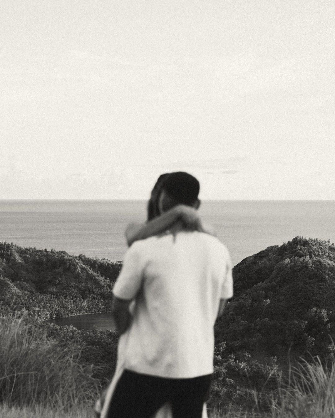 Black and white photo of a couple embracing on a coastal hilltop with ocean views.