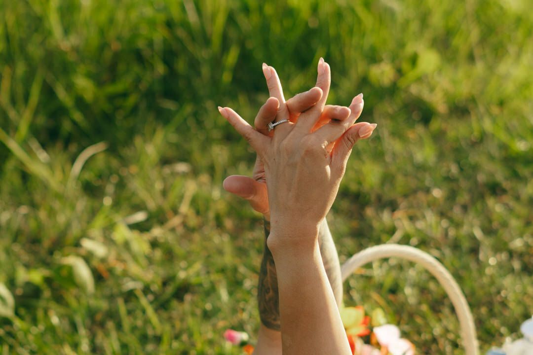 Two hands with tattoos raised against a lush green grass background outdoors.