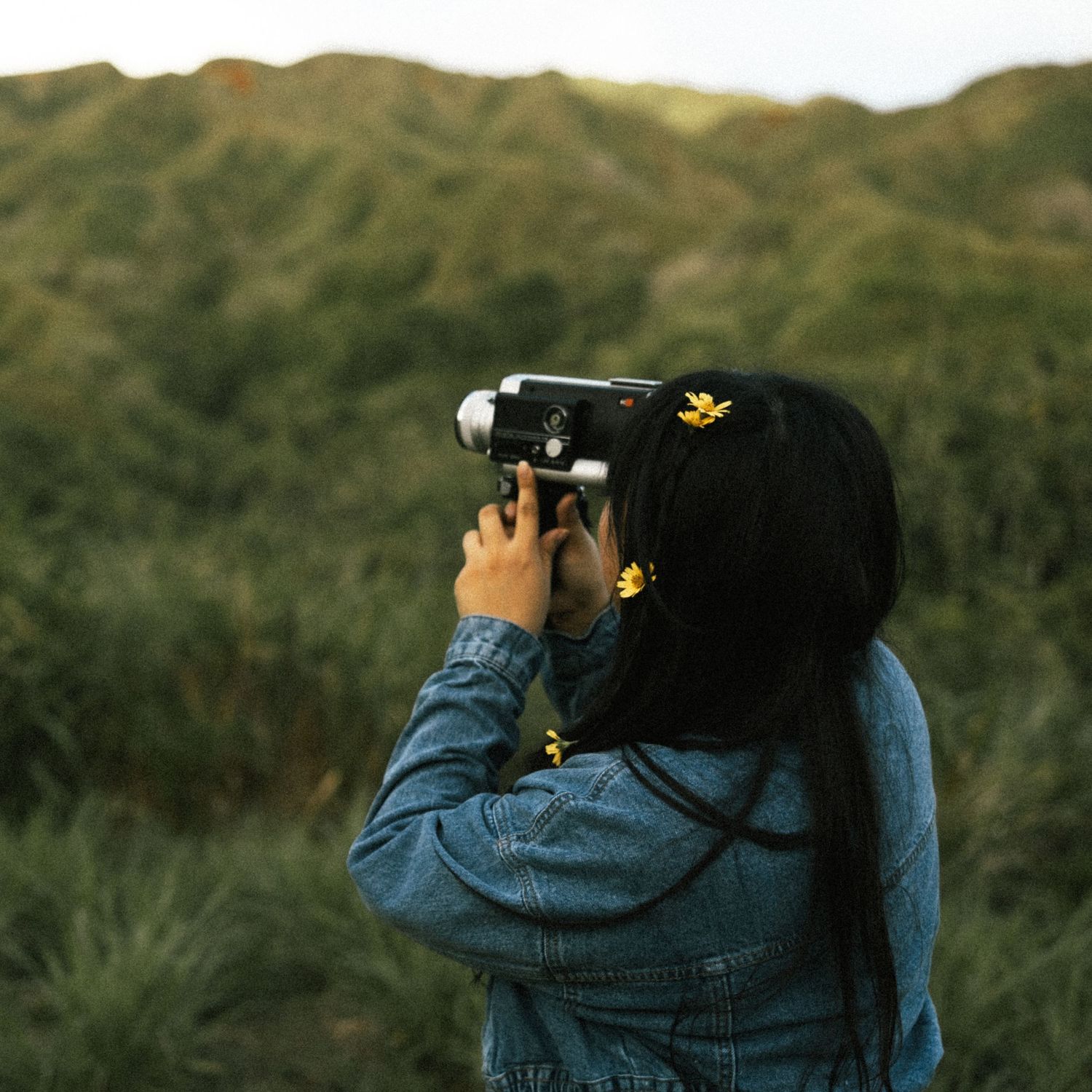 A young woman in a denim jacket photographs nature with a vintage camera, yellow hair clips accent her dark hair.