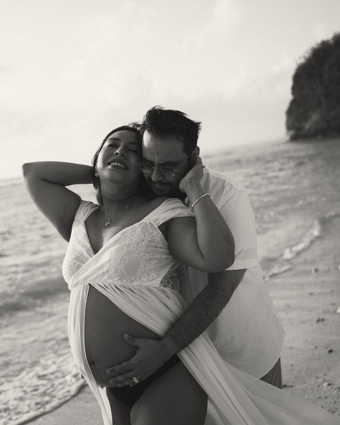 Black and white beach maternity photo of a couple embracing, woman in flowing white dress showing baby bump.