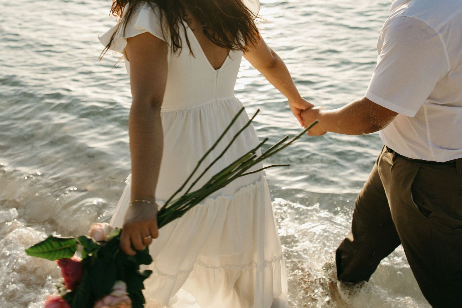 Couple holding pink roses by ocean waves, woman in white dress, romantic beach proposal scene.