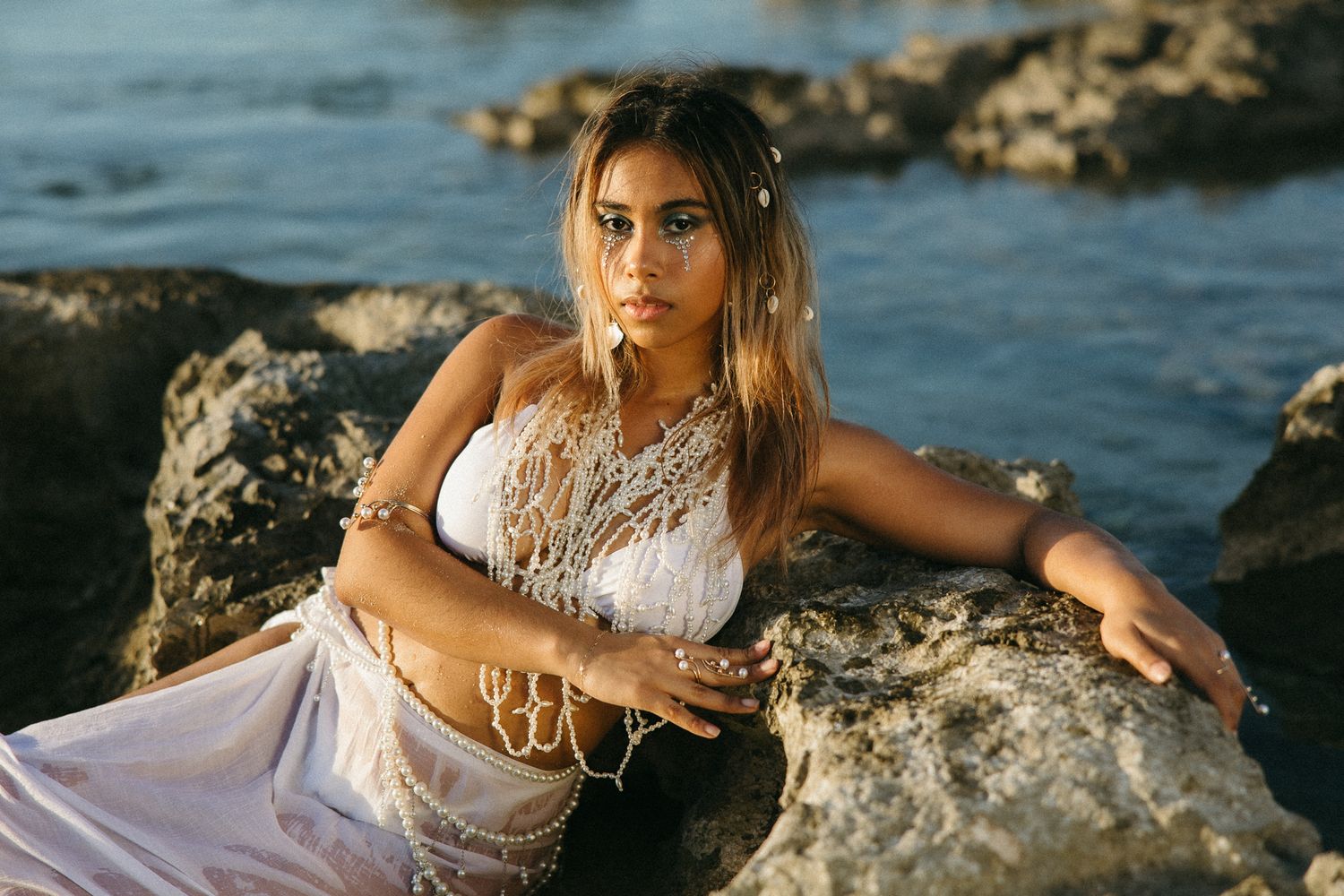 Young woman in white lace top poses on rocky shoreline with ocean backdrop in boho jewelry.