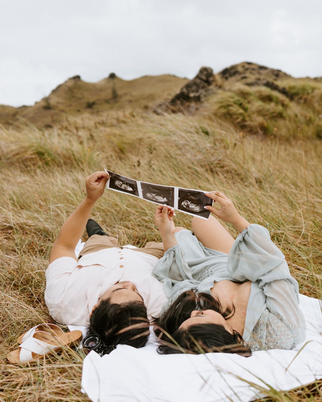 Couple lying in golden grass holding ultrasound photos, pregnancy announcement in scenic outdoor setting.