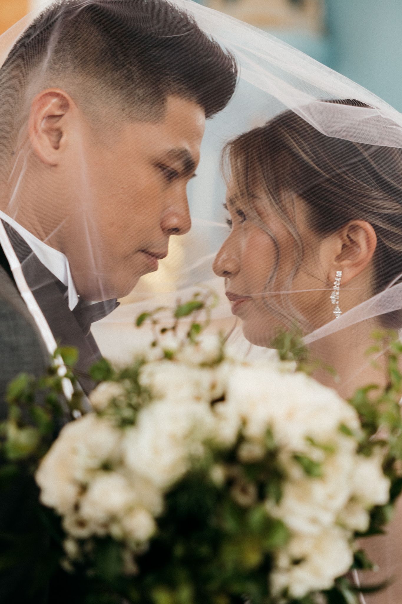 A bride and groom share an intimate moment under a wedding veil, with a white floral bouquet in the foreground.