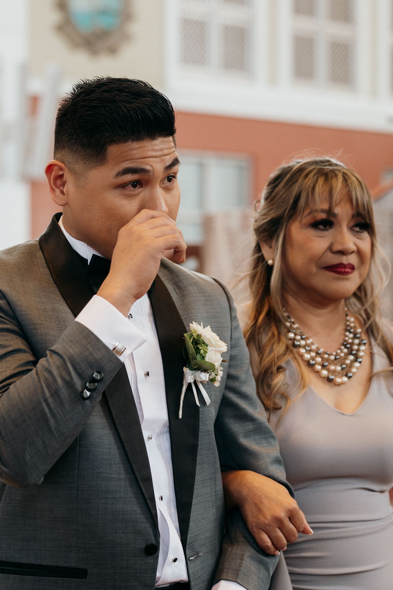 Emotional groom in gray tuxedo wiping tears at wedding ceremony, standing beside woman in pearl necklace.