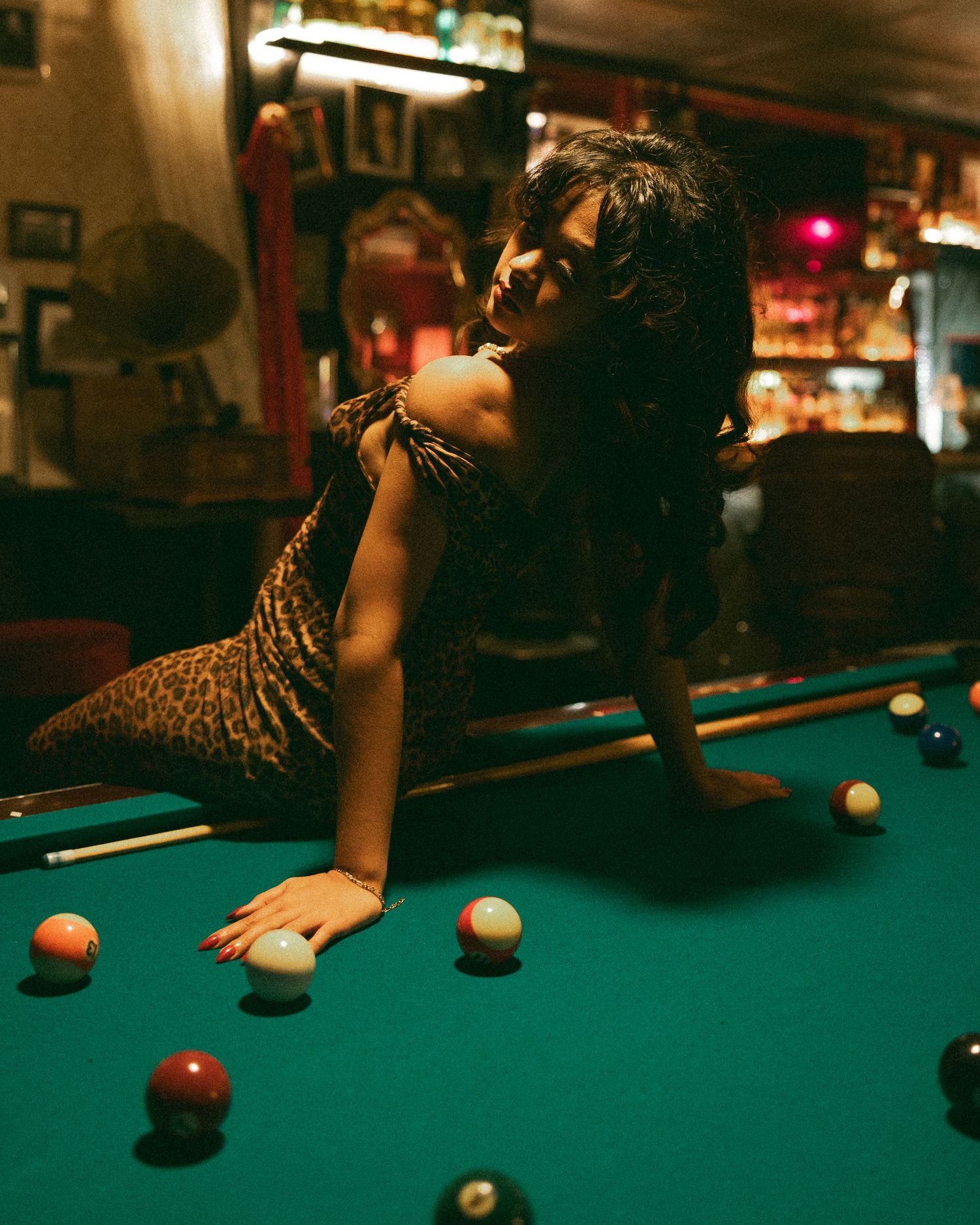 Woman in leopard print dress posing on a pool table in a dimly lit bar with billiard balls.